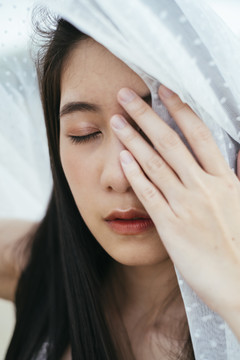 特写-年轻的亚洲长黑发女子新娘的肖像，戴着白色面纱，在户外用手遮住眼睛。