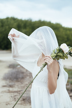 年轻的亚洲长发黑发女子新娘在户外手持白玫瑰花罩和白色面纱的肖像。