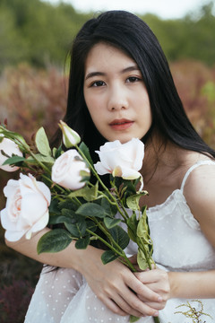 特写-年轻的亚洲长黑发女子新娘手持白玫瑰花坐在田野里的肖像。复古的。