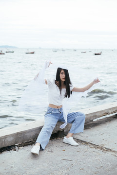 年轻的亚洲长发黑发女子戴着白纱坐在海桥上的画像。衬衫和蓝色牛仔裤长裤。