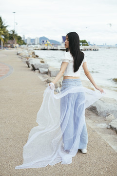 后景-亚洲时髦年轻人深色长发性感女子新娘戴着白色面纱在沙滩上散步的肖像。