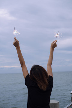 背景图：年轻的泰国亚裔女子短发举手举着两个火花头在海滩上在杜什时间。