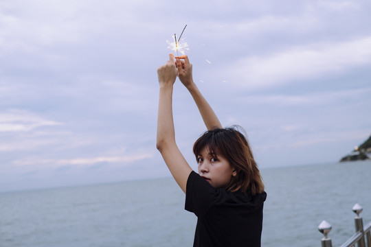 背景图：年轻的泰国亚裔女子短发举手举着两个火花头在海滩上在杜什时间。回头看摄像机。