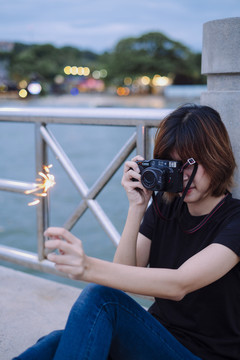 黄昏时分，一位身穿黑色衬衫和蓝色牛仔裤的泰国亚裔年轻女子坐在海边用相机拍摄闪闪发光的照片。