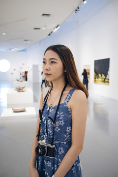 身着蓝色连衣裙的亚洲时髦美女，带着相机走进美术馆。