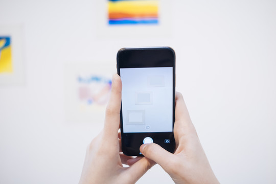 手持智能手机在白色画廊里拍摄艺术品的女人的手。