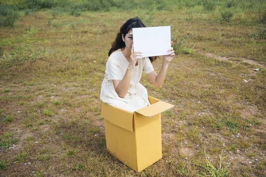 年轻的泰国亚裔女子时髦卷发白色连衣裙坐在盒子里显示空白纸的标志。复制空间。
