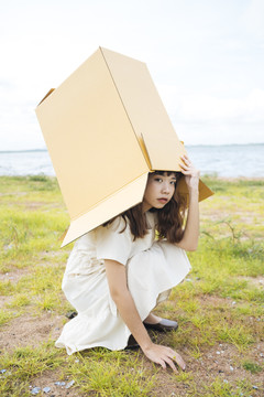 年轻的泰国亚裔女子时髦的卷发白色连衣裙把纸板箱盖在头上。坐着摆姿势。湖边可爱的女孩。