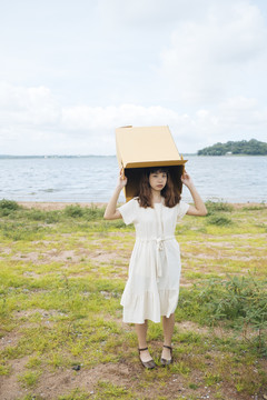年轻的泰国亚裔女子嬉皮卷发的白色连衣裙玩纸板箱放在头上在湖边。