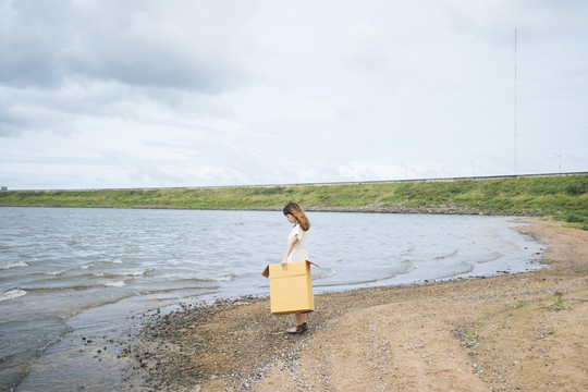 年轻的泰国亚裔女子嬉皮卷发，身穿白色连衣裙，拎着纸板箱望着湖面。
