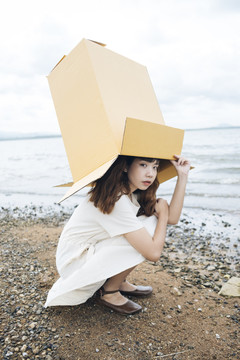 年轻的泰国亚裔女子嬉皮士卷发的侧视图白色连衣裙把纸板箱放在头上坐在湖边。