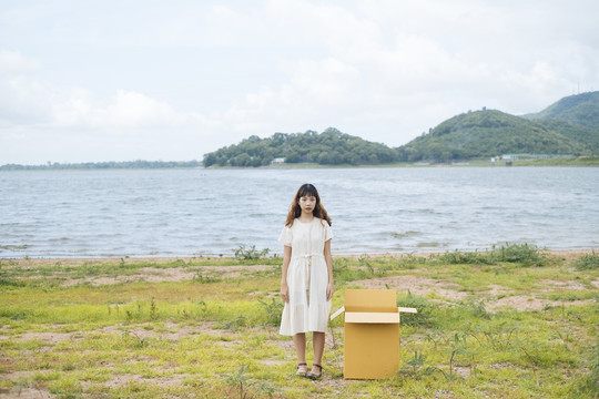 年轻的泰国亚裔女子时髦卷发，身穿白色连衣裙，手持纸板箱站在湖边。