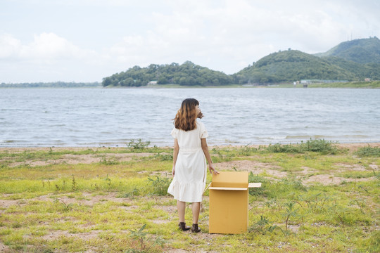 年轻的泰国亚裔女子嬉皮士卷发白色礼服站在与纸板箱在湖边。