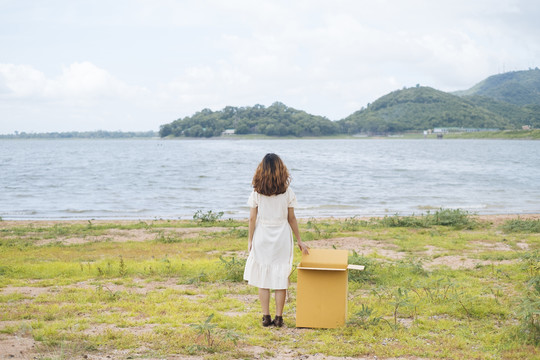 年轻的泰国亚裔女子嬉皮士卷发白色礼服站在与纸板箱在湖边。