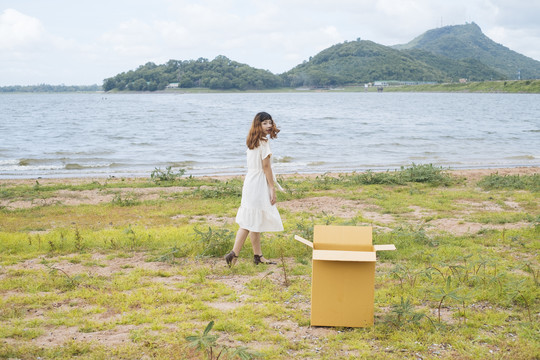 年轻的泰国亚裔女子嬉皮卷发，白色连衣裙，在湖边玩，丢下纸板箱。