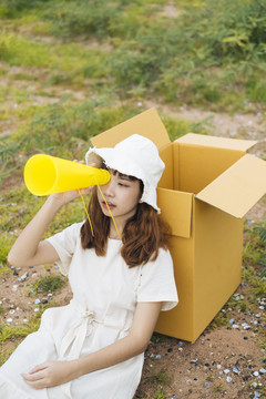年轻的泰国亚裔女子时髦卷发，身穿白色连衣裙，倚在纸板箱上，玩着玩具黄色扩音器。