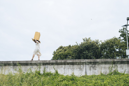 年轻的泰国亚裔女子嬉皮卷发，白色连衣裙，头戴纸板箱，走在墙上。