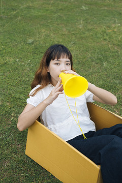 年轻的泰国亚裔女子嬉皮卷发白衬衫玩玩具黄色扬声器扩音器，坐在纸板箱在绿色领域的背景。