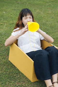 年轻的泰国亚裔女子嬉皮卷发白衬衫玩玩具黄色扬声器扩音器，坐在纸板箱在绿色领域的背景。