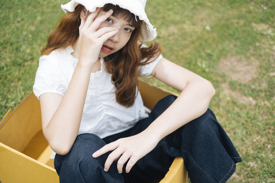 在大自然的绿色草地上，嬉皮的泰国亚裔年轻女子留着时髦的卷发，穿着白衬衫，戴着帽子，坐在纸板箱里。