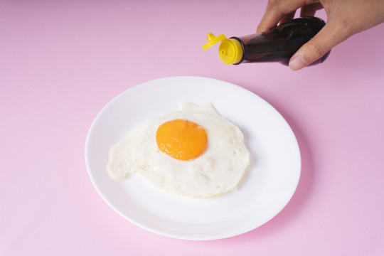 美味的煎鸡蛋放在白色盘子里，用酱油和粉红色的背景一起吃。