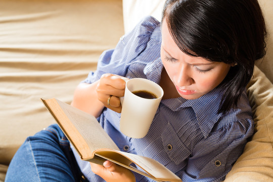 学生-年轻的亚洲妇女或女孩坐在沙发上喝着咖啡看书，她正在学习