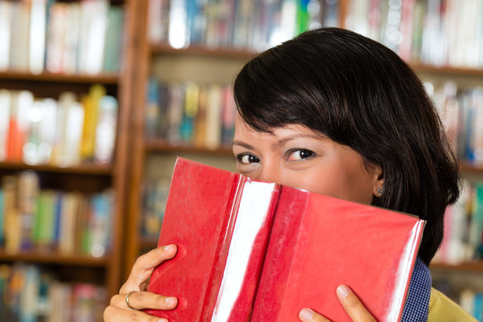 学生-年轻的亚洲妇女或女孩在图书馆学习和阅读，她躲在书后面