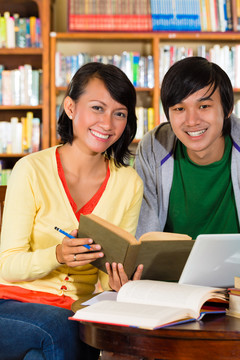 学生-年轻的亚洲妇女和男子在图书馆与笔记本电脑和书籍学习，他们是一个学习小组