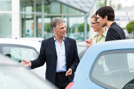 成熟的汽车经销商和年轻夫妇站在经销商的停车位前的汽车