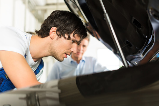 成熟的男人-客户-和年轻的机械师正在看一个带灯的引擎盖下面