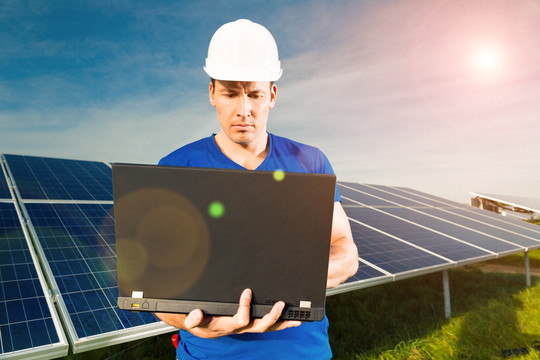 光伏系统采用太阳能电池板，通过太阳能生产可再生能源，一名技术人员手持笔记本电脑站在前面