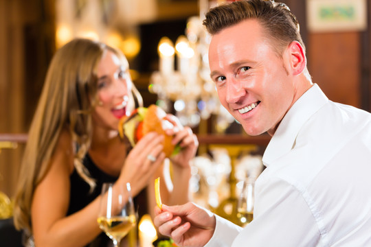 一对男女在一家高级餐厅吃快餐、汉堡和薯条，背景是一盏大吊灯