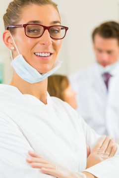 牙医在她的手术中看着观众，背景是她的同事正在给一名女性患者治疗