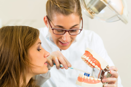 牙医在他的手术中拿着一副假牙，用牙刷向一位女性患者解释