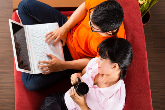 一对印度尼西亚夫妇坐在沙发上，手提电脑使用互联网收发电子邮件和网上购物
