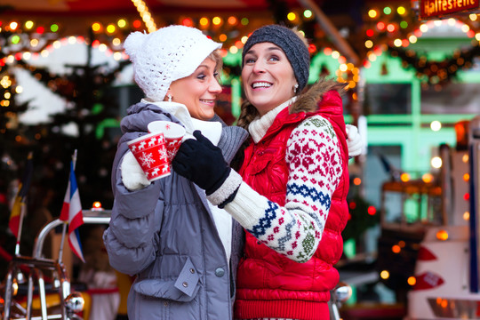 在圣诞节或圣诞节的市场上，两位女性朋友在旋转木马前喝着一杯或一杯加香料的葡萄酒或蛋奶酒或朋克酒