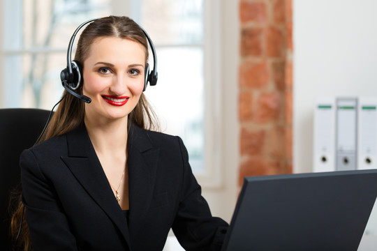 年轻的女商人或秘书在办公室工作，她坐在窗前，戴着耳机在电脑上工作，她有一个客户推销