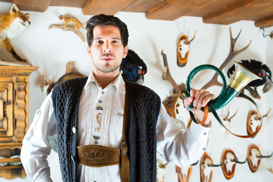 在阿尔卑斯山的小屋里，一个年轻的猎人拿着号角站在墙上，上面有不同的角、鹿角和战利品