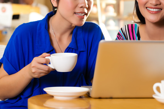 亚洲女性朋友在咖啡馆里享受休闲时光，喝咖啡或卡布奇诺，在笔记本电脑上工作