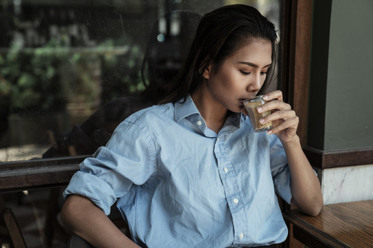 一位穿着蓝色衬衫的亚泰妇女在玻璃杯里啜饮着咖啡拿铁。