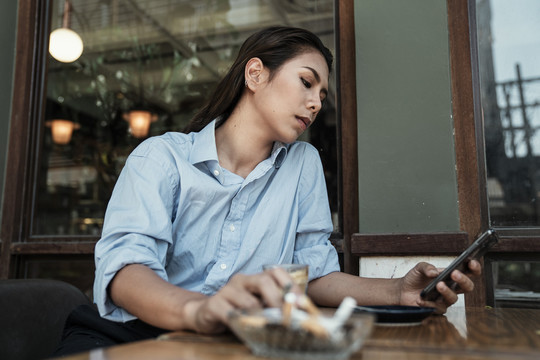 一位穿着蓝色衬衫的亚泰妇女用智能手机将香烟上的烟灰抖到烟灰缸里。