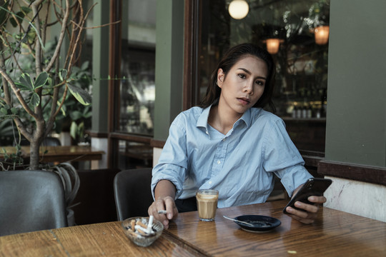 一位穿着蓝色衬衫的亚泰妇女用智能手机将香烟上的烟灰抖到烟灰缸里。