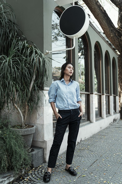 身着蓝色连衣裙的泰国亚裔女子站在街上，双手插在口袋里。