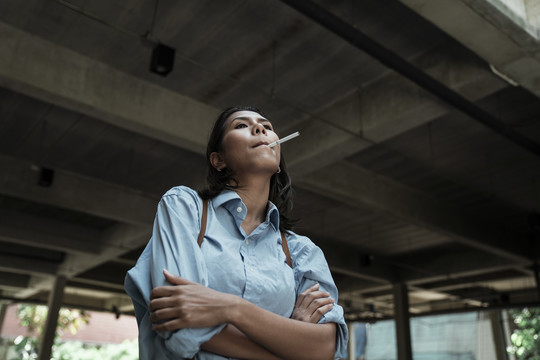 穿着蓝色衬衫的泰国亚裔女子在大楼地下室抽烟。双臂交叉，若有所思。
