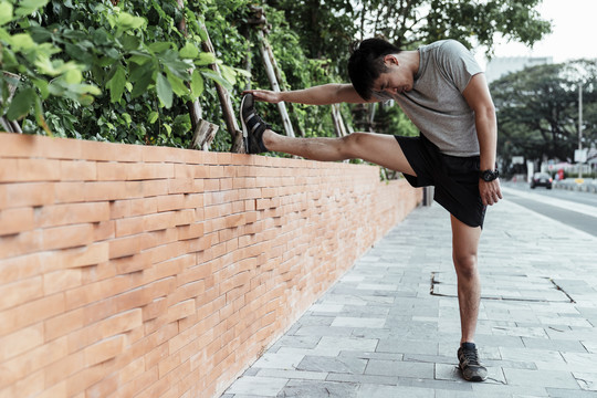 亚洲人把他的腿放在砖墙上，伸展他的腿，在街上锻炼前热身。