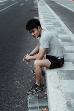 严肃的亚洲人独自坐在人行道上，看着马路。