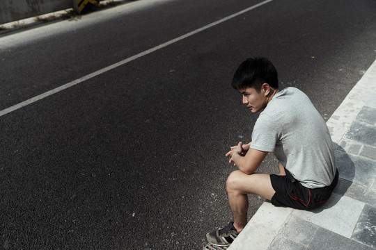 一个穿着灰色衬衫的亚洲小伙子坐在路边的人行道上。体贴的亚洲人独自坐在路边。
