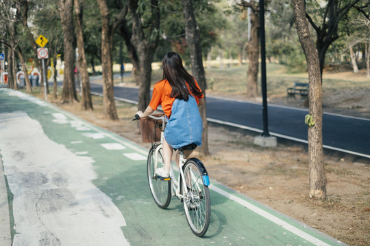 一个穿着橙色t恤和牛仔裤的长发女孩在公园的自行车道上独自骑着自行车。