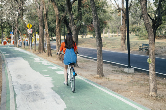 一个穿着橙色t恤和牛仔裤的长发女孩周末在公园的自行车道上骑自行车。
