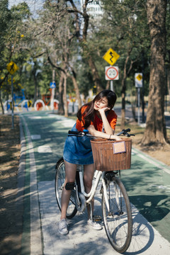 长发女孩穿着橘色t恤和牛仔裤，在公园的自行车道上停下自行车，然后把她的头放在自行车上，在阳光下闭上眼